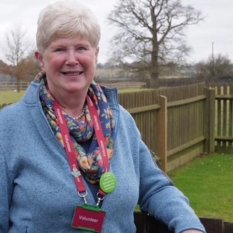 Mid shot of a happy woman leaving on a fence outside looking directly at the camera and wearing a lanyard with a badge that says ‘Volunteer’