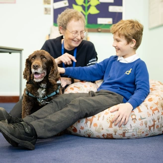 A young boy in blue uniform jumper is seated on a bean bag chair. He is stroking a brown down as a woman kneels smiling beside them