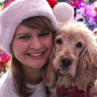 A woman wearing a Christmas santa hat, next to a light coloured dog that is wearing a bow tie. A Christmas tree can be partially seen in the background.