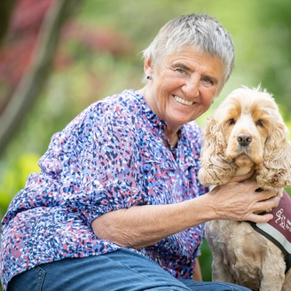 A smiling grey haird woman, kim, and beside her is Bear, a light coloured cocker spaniel hearing dog.