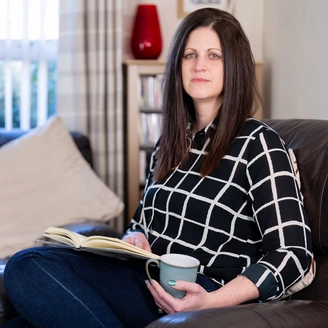 A woman with dark shoulder length hair is sitting on a dark sofa in a living room holding a book in her lap. She has a serious look on her face.