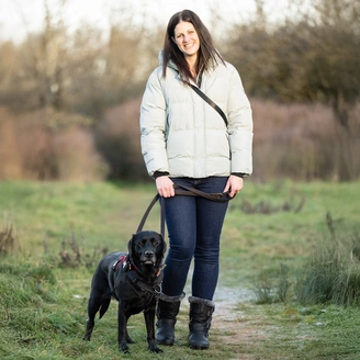 A woman wearing a white jacket standing in a field with a black dog at her side.
