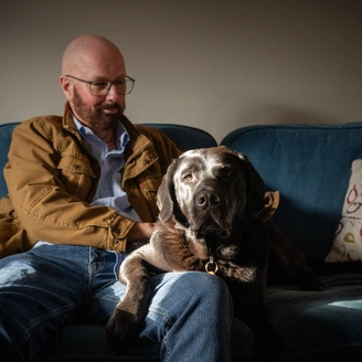 A bearded man, trevor sitting on a sofa with a large brown and white dog on his lap.
