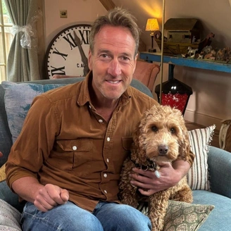 A man (ben fogle) sitting on a blue couch, his arm around a light coloured dog.