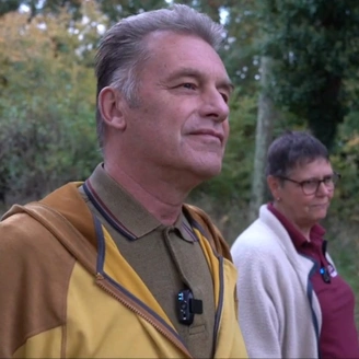 A man wearing a yellow jacket standing beside a woman in hearing dogs unform, in  a woodland setting.