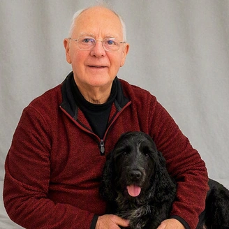 A smiling older gentleman with spectacles, sitting with a black dog resting on his lap