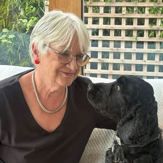A bespectacled woman with white hair smiling at a black cocker spaniel sitting on a sofa beside her