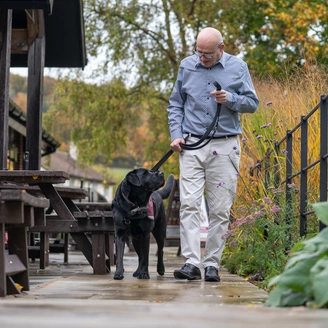 A bespectacled man walking a black labrador hearing dog on a lead. The dog is looking up at the man.