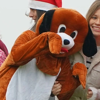 A person in a dark brown dog costume with their hands up like paws begging with smiling people in the background 