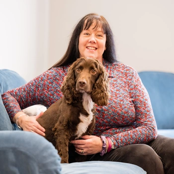 A smiling woman sat on a sofa with a brown Spaniel sat in front of her