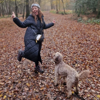 A woman, in woods, in a long coat stood on one leg with her arms in the arm looking very happy with her hearing dog stood looking up at her 