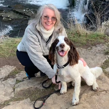 A woman crouching down but a river smiling at the camera with a happy looking Spaniel sat beside her